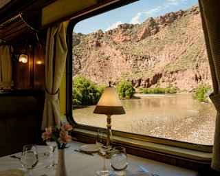 View from a table with glasses and a lamp through a window, where a slow, wide brown river cuts alongside red-stone cliffs.