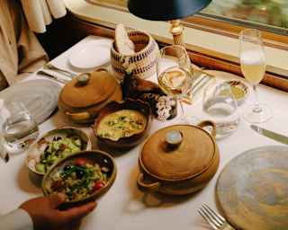 A waiter places a dish of grain salad on a table already busy with lidded pots and lunch bowls containing vibrant vegetables.
