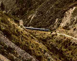 The Hiram Bingham travels on a valley track between steep forested hillsides with rocky slopes, seen from above.