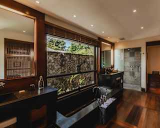 View of a villa bathroom, with floor, storage and frames in natural wood, with a long stone bath and grey-tiled shower area.