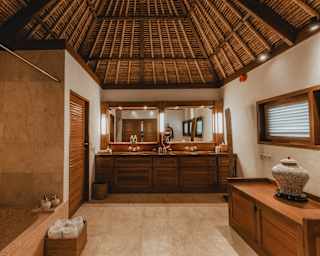 View of a high-ceilinged, serene bathroom with a stone bath, soft peach and sand tiles, and a cabinet and basin units in wood.