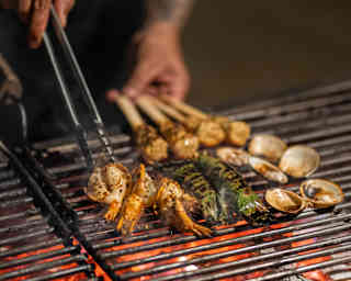 Details of a chef's hands using tongs to turn green tiger prawns, clams and fish sticks on the glowing embers of a hot grill.