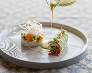 A chef's hand pours Balinese yellow paste sauce on to a plate of Mahi-mahi fish, wrapped in cucumber, with fragrant rice.