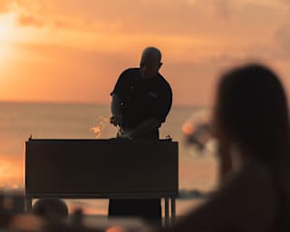 Silhouetted against the peachy sunset, a chef prepares food on a beach barbeque watched by a woman in soft-focus foreground.