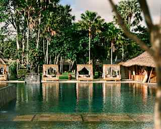 View past stone frog water spouts across a tropical aquamarine pool to a row of poolside cabanas with lush gardens behind.