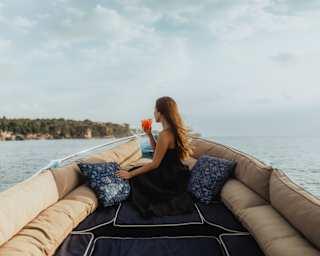 A long-haired woman in a black dress sits among cushions on the Dedari, holding an orange drink and gazing at the coast.