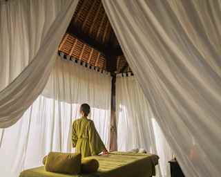 A woman in an olive-green gown stands by a treatment bed at the Beach Spa, seen from behind, through open white drapes.