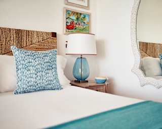 A pearl-framed mirror reflects a crisp white bed, topped with turquoise throw and blue cushion with elegant batik shell-print