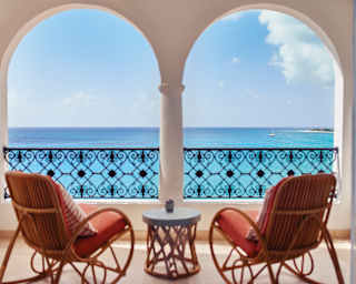 Two cane rocking chairs with terracotta cushions enjoy the view of Baie Longue through the stucco arches of a private balcony