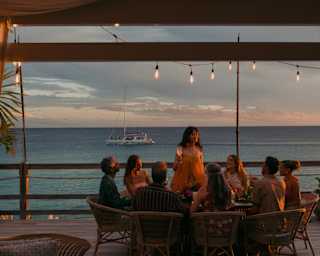 A woman stands, talking to a group of guests seated for dinner on the Laplaj deck in a view that captures a boat cruising by.