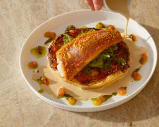 A hand pours a jug of zesty maltaise onto a dish of snapper fillet atop a papaya parmesan tart with herb and fruit garnish.