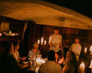 Angled image of two chefs bringing plates to a group of La Cave guests, dining by atmospheric candlelight in the cellar.