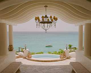 View from the cool stone lobby with undulating ceiling, chandelier and pool, out to sea where a boat floats in the blue.