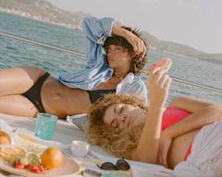 A woman with dark hair, black bikini and blue shirt and a woman with blond hair and red bikini lounge on deck with snacks.