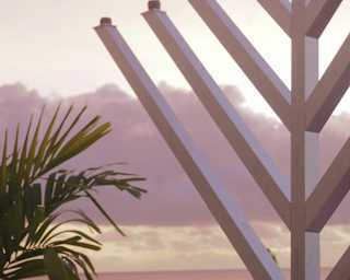 A large menorah stands outdoors at sunset with a palm tree nearby. The sky is pink and purple with scattered clouds, creating a peaceful and warm atmosphere.