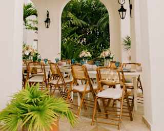 View through a wall arch of a long celebration table with cushioned cane chairs, laid with flowers and blue glassware.