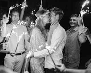 Monochrome image of a blonde bride in a flower headband smiling as she kisses her groom, circled by friends waving sparklers.