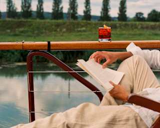 A person in light clothing reads a book whilst reclining on a deckchair by a lake; a red drink rests on the wooden railing, with trees and a cloudy sky in the background.