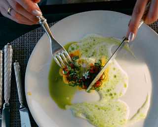 A person in a white top uses a fork and knife to cut into a gourmet dish with green sauce, served on a white plate on a woven placemat with silver cutlery.