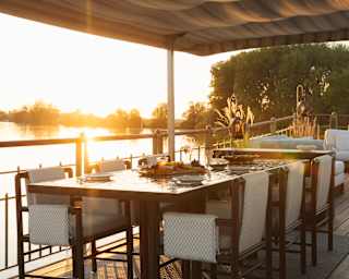 A long dining table with chairs is set on a decking by the water at sunset, under a wavy fabric canopy.