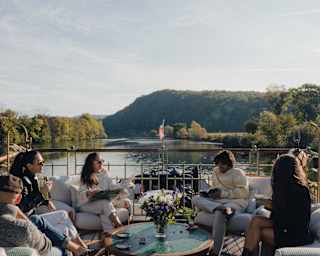 Five people sit and talk around a circular table with drinks on a boat deck, surrounded by water and trees, with a mountain visible in the background under a clear sky.