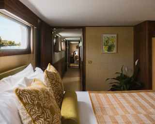 A hotel room with a bed featuring patterned yellow cushions and a striped throw, looking through a doorway into a connecting room.