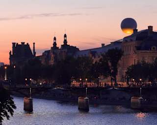 A sunset view of a river in Paris with silhouetted historic buildings, a bridge, and a large art installation resembling a white sphere on a rooftop, softly lit against the evening sky.