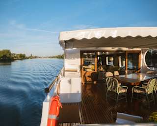 The covered deck of a boat with a dining table and chairs, moving along a calm river.