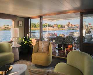 Interior of a luxury boat lounge with green and yellow seating, opening onto a deck with a dining table overlooking a canal and village.