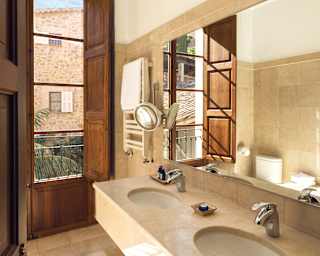 Green palms and ancient stone walls can be seen through the full height wooden shutters of marble bathroom with twin vanity