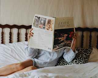 A woman in pyjamas lies on a bed with a breakfast tray and a copy of La Resi Tribune, which she holds in front of her face.