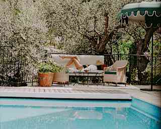 A woman lies on a garden sofa with a hat on her face, seen across a pool with a backdrop of olive trees and craggy hills.