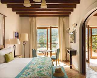 Looking across a crisp white bed with turquoise floral throw is a view of a private pool accessible via open French doors