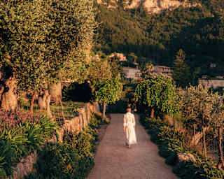 A woman in a white dress walks along a path between gardens held back by stone walls and borders, seen in golden light.
