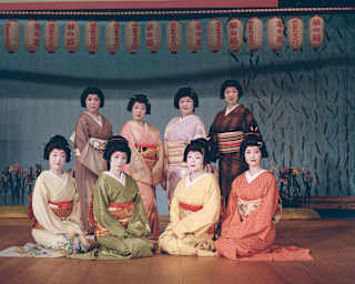 Eight women in traditional pastel Japanese kimonos and pale make-up pose below a row of lanterns, photographed by Cecy Young.