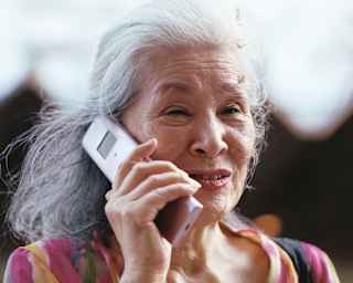 An older Japanese woman with grey hair, minimal make-up and a pink-patterned top chats on a mobile phone, seen in close-up.