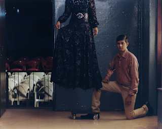 A man in red-striped shirt and beige trousers kneels by a woman in a black lace dress, apparently floating above the floor.