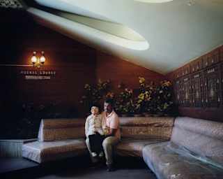 An Indian ventriloquist in a pink shirt and sage trousers sits on a large, curved banquette with his dummy on his knee.