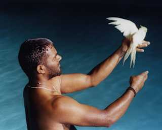 Illusionist Nikhil Raj stands bare-chested in water with a white budgerigar sitting on his outstretched hand, seen from above.