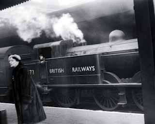 Historic image of a British Railways stream train passing by a platform, where a woman in a long fur coat and white beret walks.