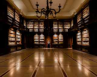 A grand, dimly lit library with multi-level wooden bookshelves lining the walls, a central chandelier, and a tiled floor.