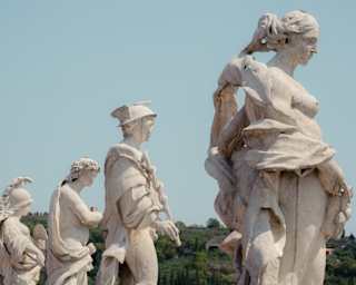 Four classical marble statues of figures standing in a line against a clear blue sky.