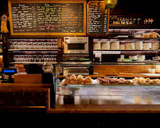 Bar counter with glass cases displaying various small appetisers, with menus written on blackboards hanging on the wall behind shelves of wine bottles and crockery.