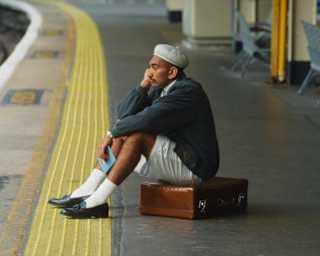 A man in a jacket, shorts, and a beret sits on a suitcase at a railway station platform, resting his chin on his hand and looking thoughtful. Empty benches and yellow tactile paving are visible nearby.