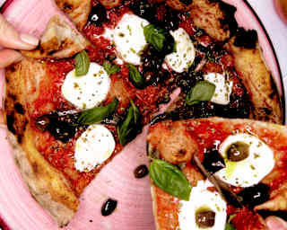 Diners take slices of wood-fired pizza with tomato sauce, fresh mozzarella, basil laves and olives from a pink plate.
