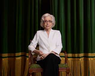 Fernanda Montenegro, with grey hair, glasses, dark trousers and white top, sits elegantly on a chair on stage, looking ahead.