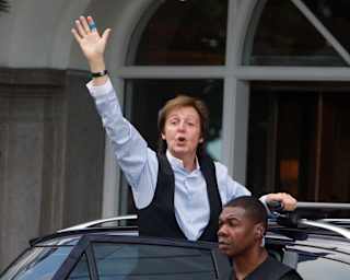 A guard stands by as Sir Paul McCartney in a blue shirt and black waistcoat waves from the open car door outside the hotel.