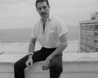 Black and white photo of Freddie Mercury sitting on a roof terrace wall in black pants and white shirt, holding a cigarette.