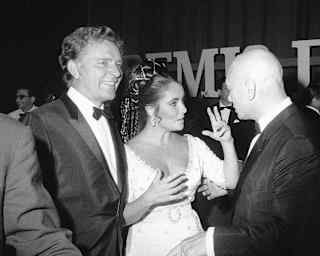 With Richard Burton at her side, Elizabeth Taylor, in a white dress and braided hair, talks to a guest, in black and white.