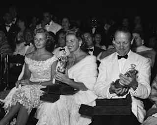 Ingrid Bergman, wearing white fur, and Sir Ashley Clarke hold their awards at the Taormina Film Festival, in black and white.